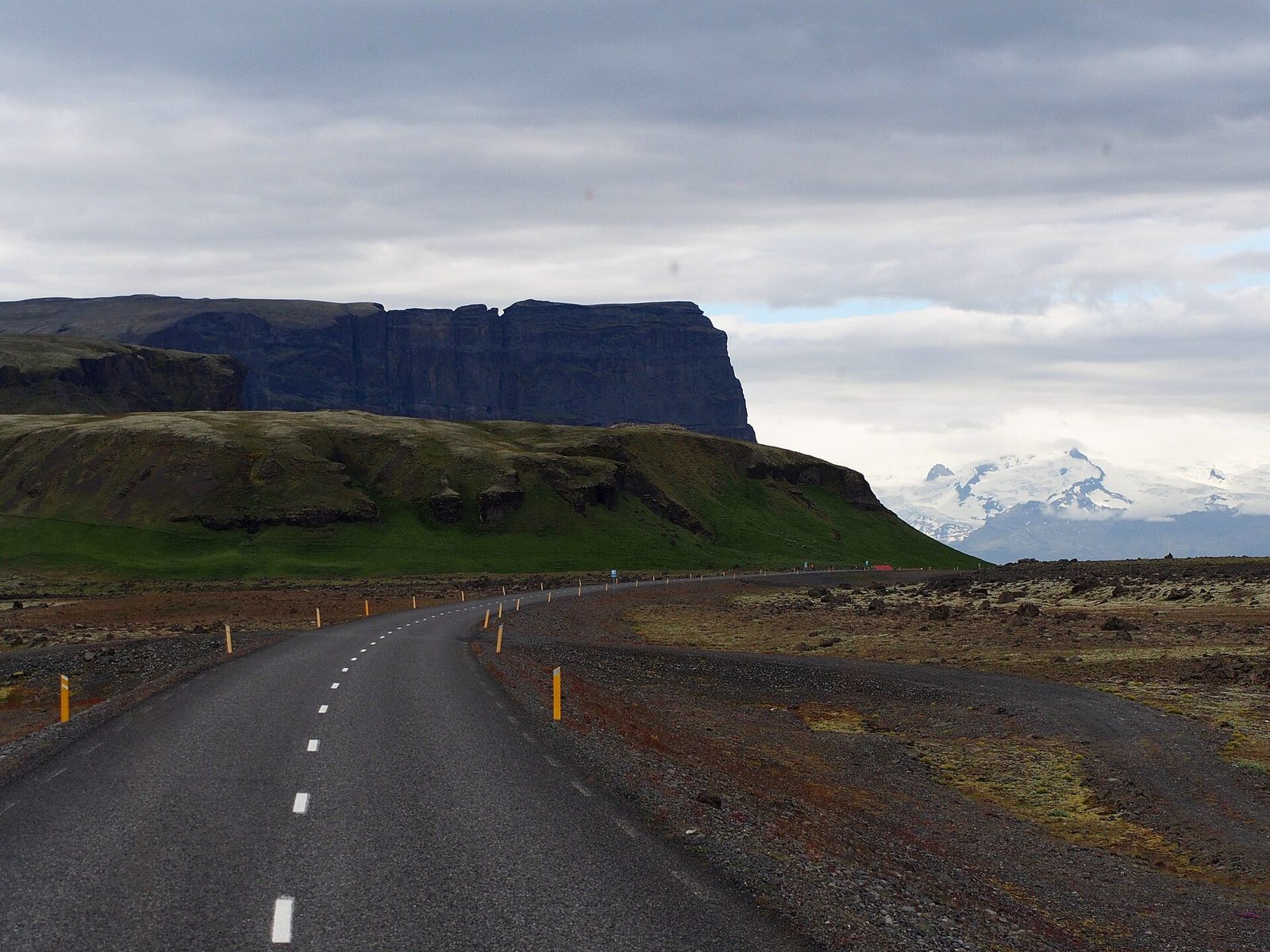 An empty stretch of Iceland's Ring Road (Route 1) curving beneath the Lómagnúpur cliffs in South Iceland, with Öræfajökull in the distance