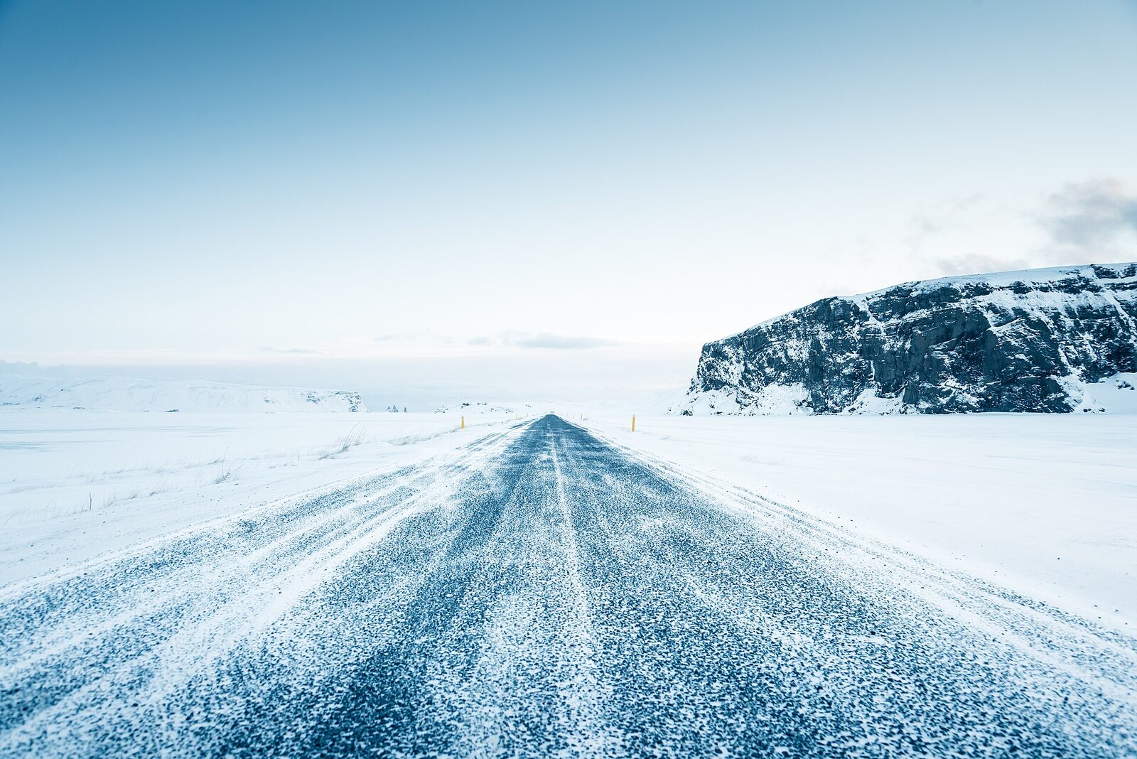 Drifting snow blowing across an empty paved road in Iceland, with marker posts and snow-covered ground either side
